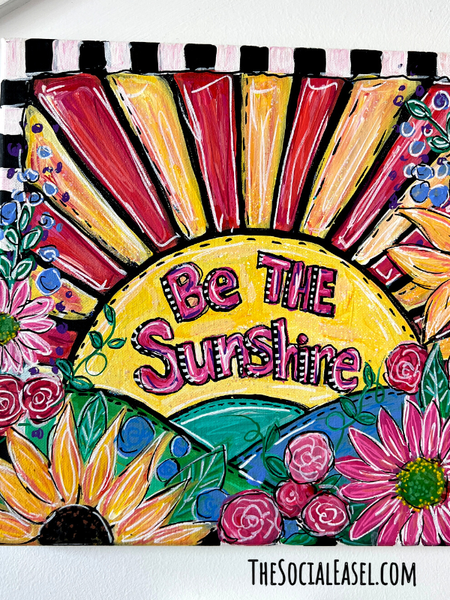 Be The Sunshine painted in bright colors of pink accented with white, black and blue. A Sun design with flowers on the bottom of the canvas. 