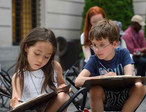 A young girl aged around 6 with long, brown hair and wearing a white summer top, and a young boy of similar age, wearing spectacles and a blue t-shirt are sitting on chairs with easels on The Met plaza. Both are sketching on paper with a pencil. Adults are seated behind in the distance, also drawing and sketching.