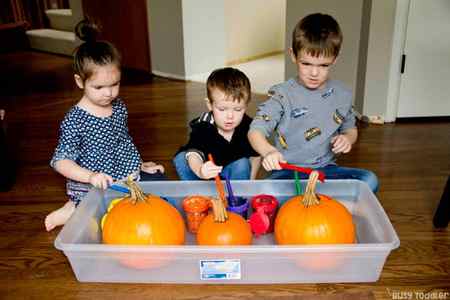 Wide photo of 3 children beginning to paint large orange pumpkins. Their brushes are up and paint is dripping off.