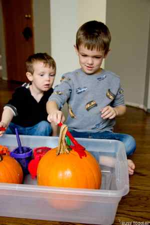 A toddler looks over the shoulder of a 5 year old, who is applying red paint to an orange pumpkin.