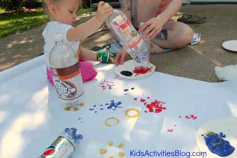 Step 5 - Paint Flowers with Water Bottles -Add paint to a paper plate, and dip a water bottle into it- little girl stamping paint and water bottle on a large sheet of paper to make flowers- blue flowers, yellow flowers, red flowers