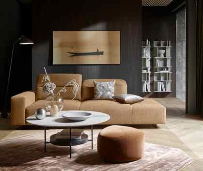 A spacious living room with a mustard-colored sofa, a ceramic coffee table, a camel cotton footstool in the front, and a grey wall system in the back.