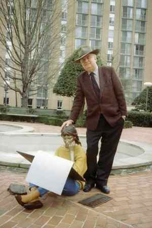 Seward Johnson rests his hand on the head of a statue of a girl sitting on the ground with a large sketch pad