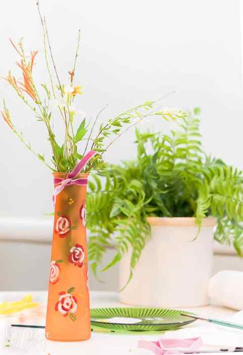 A beautiful Hand Painted Floral Dollar Store glass orange Vase with acrylic paint. A few pieces of sage are in the small vase with velvet ribbon tied around the top. A fern in a light pink planter is behind it. 