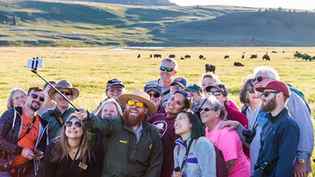 Ranger taking a selfie with a group of people with a herd of bison in the distance behind them